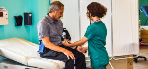 Young female student standing while taking blood pressure of older man's left arm. Man is sitting on the edge of an examination table.