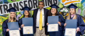 Bill Strickland surrounded by 4 females each holding a program diploma. The females are dressed in cap and gowns. Everyone is smiling.